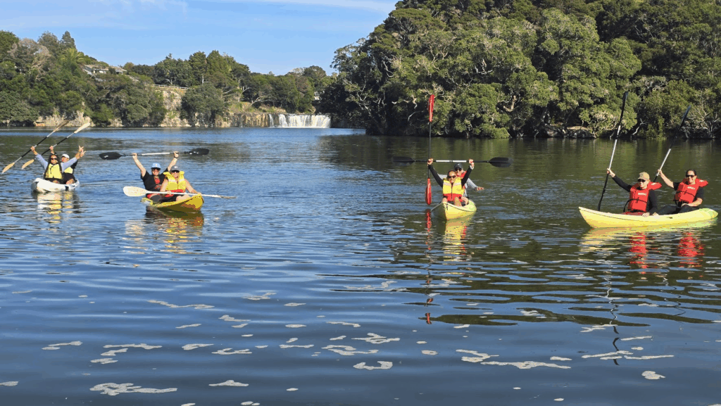 People in four 2-perosn canoes hold up paddles and their arms. Behind them, a waterfall is visible. They appear to be in an inlet, with trees and vegetation visible on both sides.