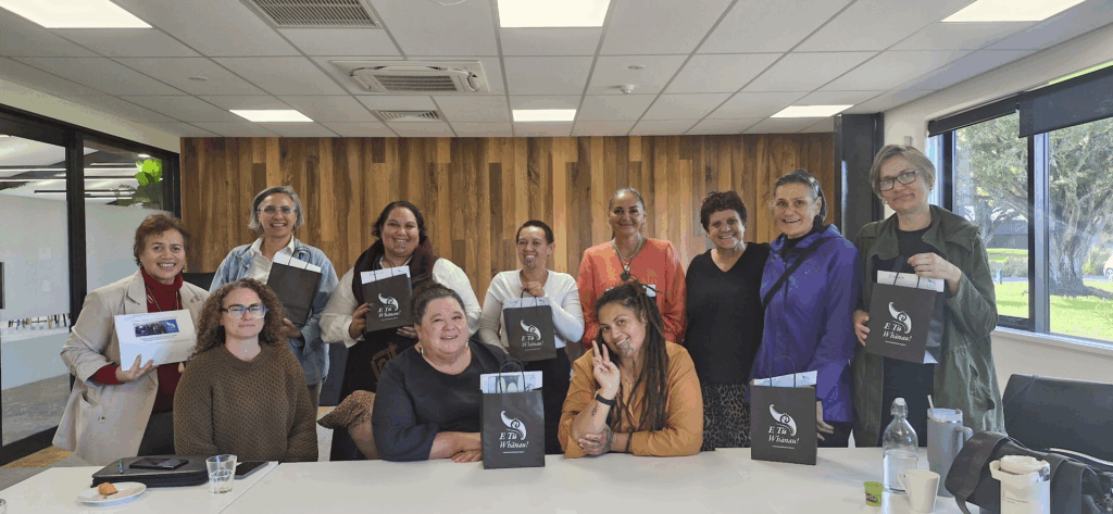 A group of 11 Mana Wāhine Collective women sit or stand together along a table. They are holding black E Tū Whānau paper bags.