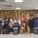 A group of 11 Mana Wāhine Collective women sit or stand together along a table. They are holding black E Tū Whānau paper bags.