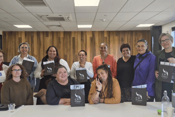 A group of 11 Mana Wāhine Collective women sit or stand together along a table. They are holding black E Tū Whānau paper bags.