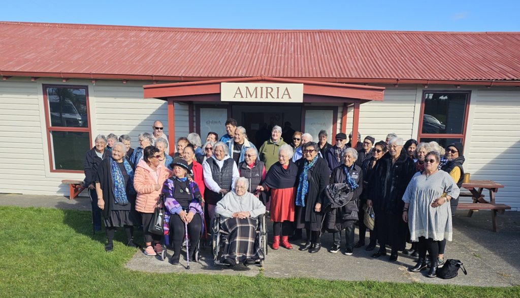 A group of about 30 kaumātua from Nannies Against P are pictured before a building named 'Amira" that has a red roof. Two of the kui are int wheel chairs.