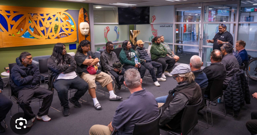 A group of people are pictured at a Tamaiti Whāngai wānanga. About 15 people can be seen seated in two rows facing each other. At the far end, a man is standing talking to the group. On the wall a koru patterned wood carving is visible.