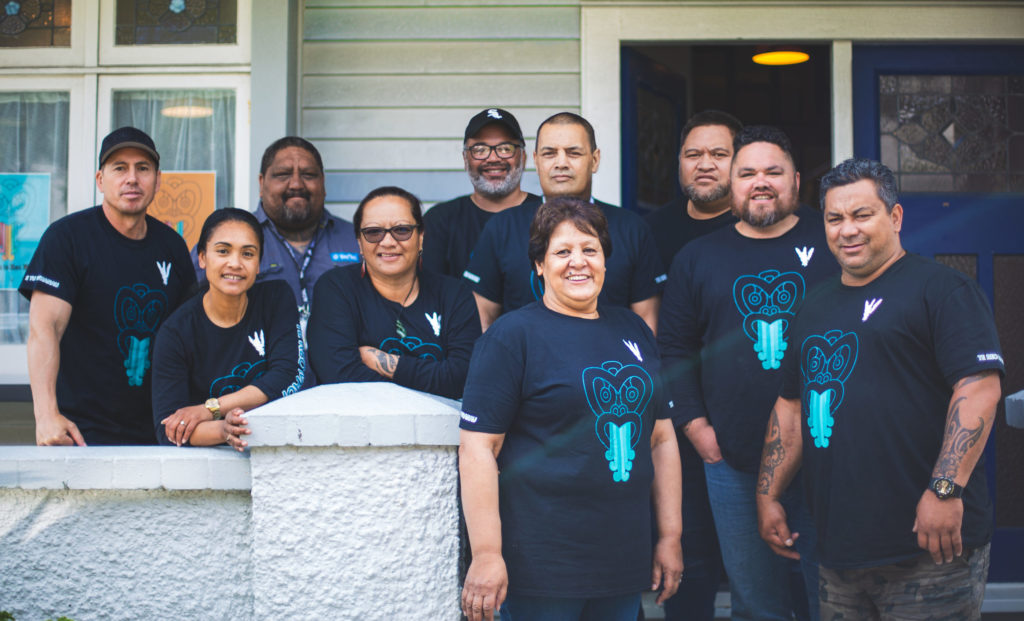 A group of Tamaiti Whāngai kaimahi are pictured together outside a building. The all wear t shirts with the Tamaiti Whāngai logo. Of the 10 people, three are wāhine.