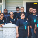 A group of Tamaiti Whāngai kaimahi are pictured together outside a building. The all wear t shirts with the Tamaiti Whāngai logo. Of the 10 people, three are wāhine.