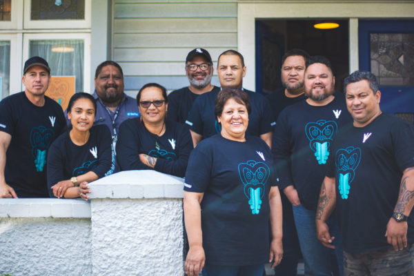 A group of Tamaiti Whāngai kaimahi are pictured together outside a building. The all wear t shirts with the Tamaiti Whāngai logo. Of the 10 people, three are wāhine.