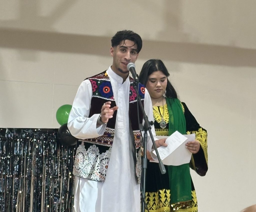 Two NFACT youth leaders, Zaki Ayubi and Aseya Rezai,, are dressed in colourful traditional costume from Afghanistan. They are standing at a microphone MCing an event.