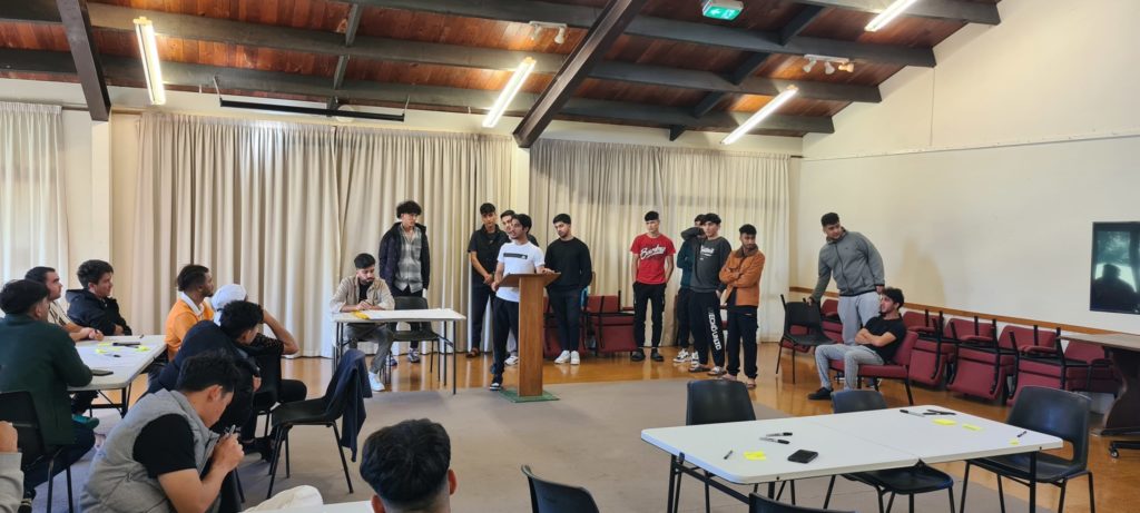 Zaki stands at a lectern addressing a room of about 20 young men in a workshop.
