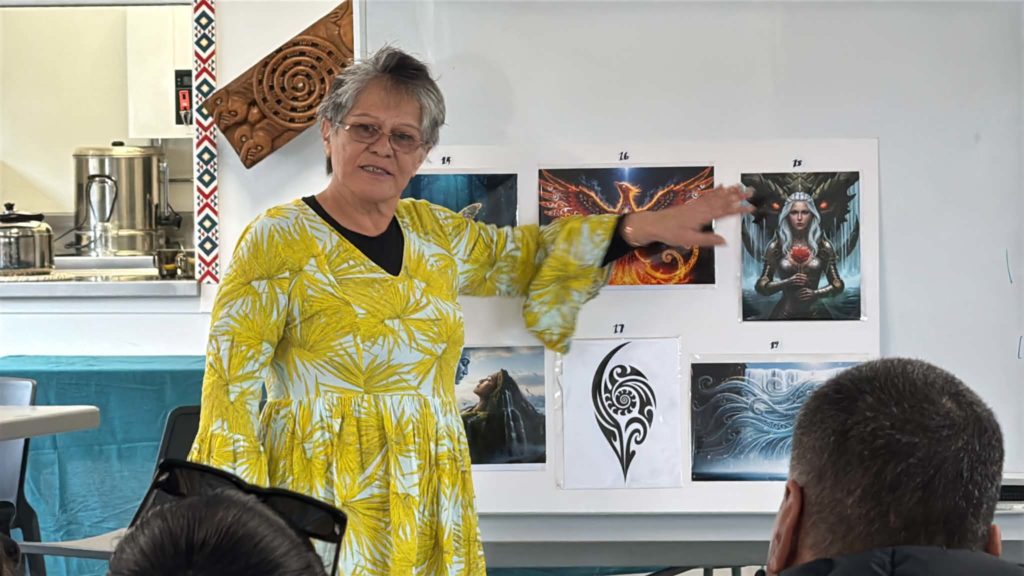 A woman is pictured wearing a bright yellow dress. She gestures towards a board behind her, on which several images made as part of Atamai Ora wānanga are visible. These images are of waves, a woman holding a styalised heart while a demon looms close behind her, a burning phoenix. Other images are not discernible. The backs of two people's heads are visible - the woman in yellow is speaking