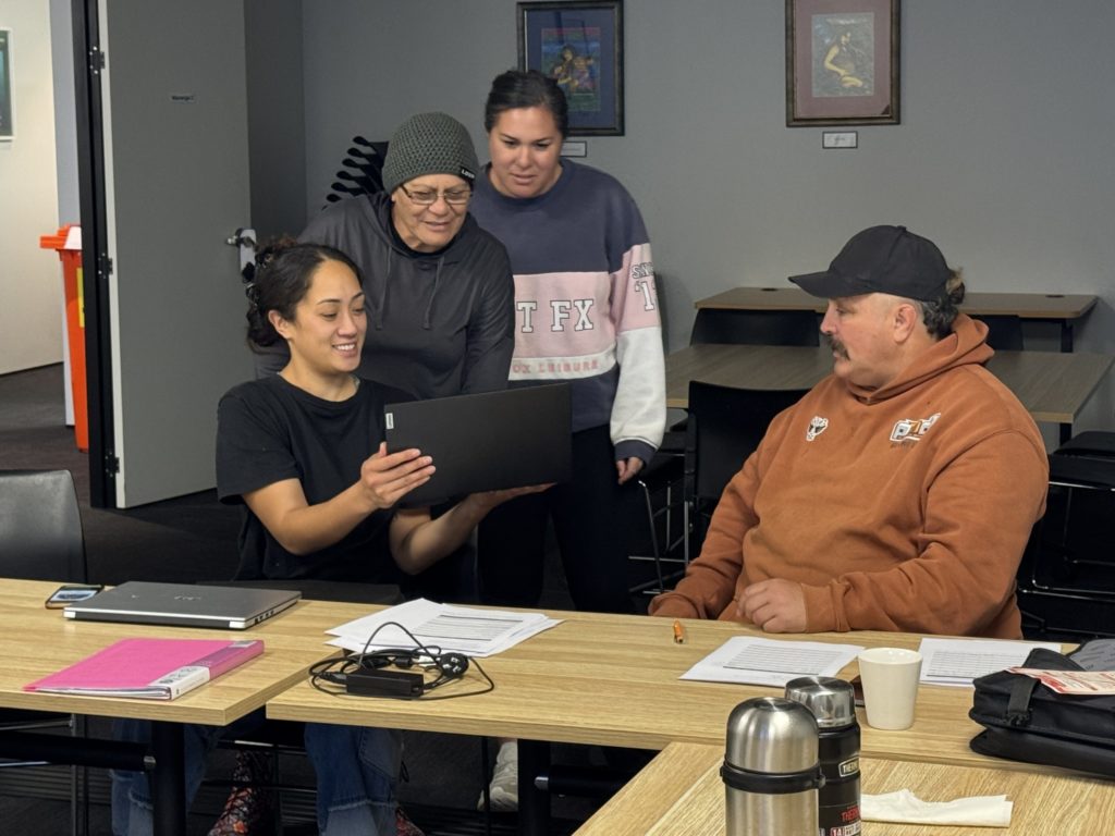 Four Atamai Ora wānanga participants are pictured together. Three women of various ages look at something on a laptop while a man looks on. In front of them, various papers, pens and other learning equipment are visible.