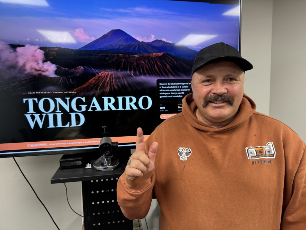 A middle aged man smiles and points a finger up in the air. Behind him, a large screen displays the words "Tongariro Wild" over the top of a picturesque landscape of two volcanoes, craters and hills. This is the image of a website he built through Atamai Ora wānanga.