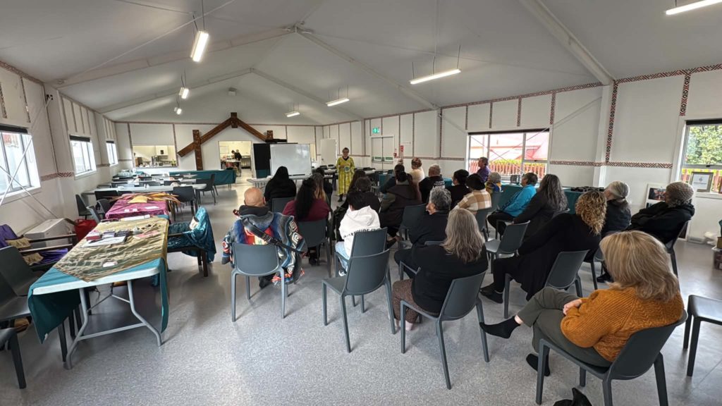 Lots of people sit in rows while a woman in a yellow dress speaks to them at an Atamai Ora wānanga. They are seated inside.