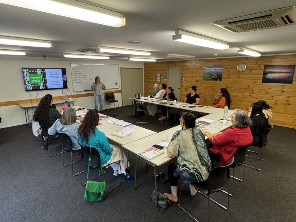 People sit in around desks arranged in u-shape at the end of which is a whiteboard and large TV screen. They all have various pens. paper and booklets with them. A woman stands in front of the whiteboard, addressing the people attending the Atamai Ora wānanga.