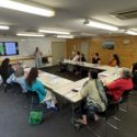 People sit in around desks arranged in u-shape at the end of which is a whiteboard and large TV screen. They all have various pens. paper and booklets with them. A woman stands in front of the whiteboard, addressing the people attending the Atamai Ora wānanga.