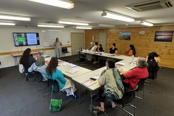 People sit in around desks arranged in u-shape at the end of which is a whiteboard and large TV screen. They all have various pens. paper and booklets with them. A woman stands in front of the whiteboard, addressing the people attending the Atamai Ora wānanga.